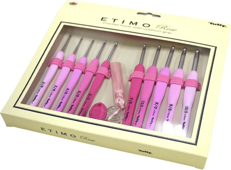 Op de afbeelding: Een set Etimo Rose haaknaalden met kussengrepen, in een cr&egrave;mekleurige doos. De haaknaalden zijn in verschillende roze tinten, met maten van 2/0 tot 10/0. De doos bevat een roze kwast en een klein roze accessoire.
