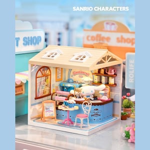 Op de afbeelding: Een miniatuur Sanrio Cinnamoroll Cafe speelset. Het café heeft een blauwe toonbank, een kleine tafel met een taart en een roze stoel. Het dak is beige met twee kleine ramen. Het bord zegt "Cinnamoroll Cafe".