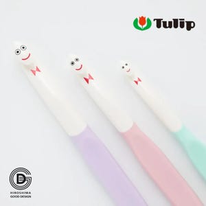Puede incluir: Tres bolígrafos caprichosos con caras sonrientes y mangos pastel en lila, rosa y verde menta. Cada bolígrafo tiene un cuerpo blanco y una pajarita roja. El logotipo de Tulip es visible. Los bolígrafos están diseñados con un tema de dibujos animados.