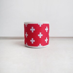May include: A cylindrical roll of elastic fabric featuring a vibrant red background with a repeating pattern of small white plus signs. The material has a textured, woven appearance with clean, finished edges at the top and bottom of the roll.