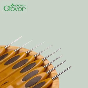 Puede incluir: Un conjunto de ganchillos con mangos marrones y puntas de metal plateado. Los ganchillos están colocados sobre una superficie de madera, con el logotipo de Clover. Los mangos tienen un diseño ergonómico.