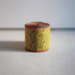 May include: A cylindrical roll of woven webbing tape featuring a vibrant yellow and black tiger stripe pattern. The edges of the roll are finished with a thin, solid red border, and the top surface shows the tightly coiled brown interior layers.
