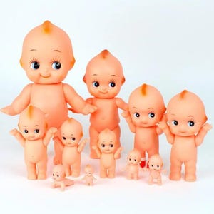 May include: A group of peach-toned baby dolls with large eyes and small orange hair tufts. The dolls are in different sizes and poses, some standing with arms outstretched, others kneeling.