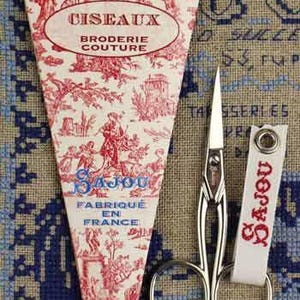 以下が含まれることがあります： 赤と白の模様のケースに入ったSajou刺繍はさみ。ケースにはフランス語のテキストとイラストが描かれています。銀色のハサミには、白いSajouタグが付いています。