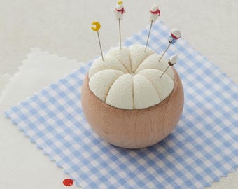 May include: A round, light brown pincushion with a cream-coloured, segmented top. Several decorative pins with small character toppers are inserted into the cushion. The pincushion sits on a blue and white checked fabric.