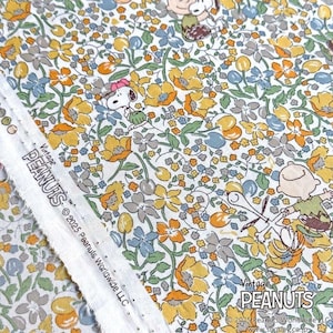 Pode incluir: Tecido floral com personagens Peanuts. O design apresenta Snoopy e seus amigos entre flores amarelas, laranja, azuis e cinzentas sobre um fundo branco. O texto "Vintage PEANUTS" é visível.