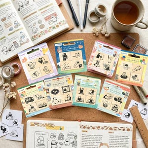 May include: A collection of Japanese stamps featuring cute cartoon characters and scenes. The stamps are packaged in colorful cardboard with the word "STAMP" visible. The image also includes a journal, washi tape, and a cup of tea.
