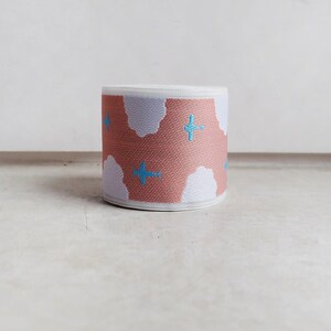 May include: A roll of elastic fabric featuring a repeating pattern of light blue airplanes flying across a dusty rose background with white cloud shapes. The woven material has a textured surface and a consistent width throughout the entire roll.