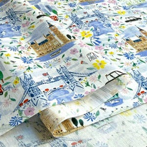 May include: White fabric with a repeating pattern of London landmarks, including Big Ben and Tower Bridge, interspersed with colorful flowers. The design features a watercolor style with blue, yellow, pink, and green accents. The fabric is folded, showing the pattern's continuity.