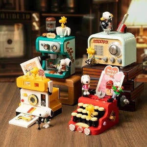 Pode incluir: Uma coleção de modelos em miniatura com o tema Snoopy, incluindo uma máquina de café, um rádio, uma câmara e uma máquina de escrever. Os modelos são de várias cores, como verde-azulado, amarelo e vermelho, e apresentam as personagens Snoopy e Woodstock. A máquina de escrever tem o texto "You're a Catch!"