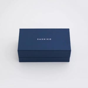 May include: A rectangular, dark navy blue box with a lid. The word SASHIKO is printed in white, sans-serif capital letters centered on the top surface of the lid. The box has a clean, solid-colored finish and a simple, structured design.