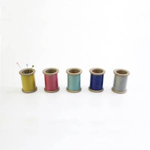 Puede incluir: Cinco carretes de hilo de madera, cada uno con un color de hilo diferente. Los carretes están dispuestos en una fila de izquierda a derecha: amarillo, rosa, azul claro, azul oscuro y gris.