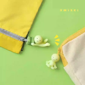 Puede incluir: Primer plano de una bolsa amarilla con cremallera plateada y una pequeña figura humanoide brillante. La figura es de color verde claro y tiene una cara sencilla. La palabra "SMISKI" está impresa en blanco sobre el fondo verde.