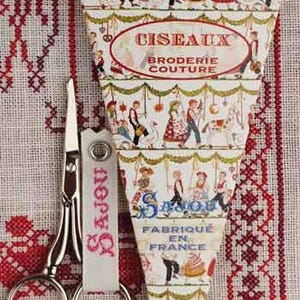Op de afbeelding: Zilveren schaar met een witte leren handgreep, met het woord "Sajou" in roze schrift, ligt op rood en wit geborduurde stof. Een decoratieve papieren schede met de tekst "Ciseaux Broderie Couture" is ook aanwezig.