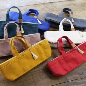Op de afbeelding: Een verzameling canvas draagtassen in diverse kleuren, waaronder blauw, groen, geel en rood. Elke tas heeft een handvat, een kleine leren patch en een decoratieve kwast. De tassen zijn geplaatst op een houten oppervlak.