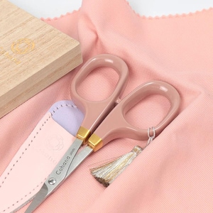 Pode incluir: Tesoura rosa com detalhes dourados e uma borla, sobre um tecido rosa. A tesoura está em uma bainha de couro rosa claro. Uma caixa de madeira está no fundo. O texto "Cohana Japan" é visível na tesoura.