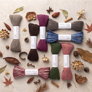 May include: A collection of eight skeins of Sashiko thread, each labelled 100m, in various colours including beige, dark brown, purple, grey, blue, olive green, dusty pink, and a variegated pink and white, arranged with scattered dried botanicals.