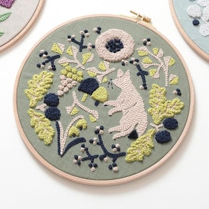 May include: Embroidered hoop art featuring a light pink cat surrounded by green, yellow, and navy blue foliage on a sage green background. The design includes a large flower and various plant elements, all within a wooden hoop.