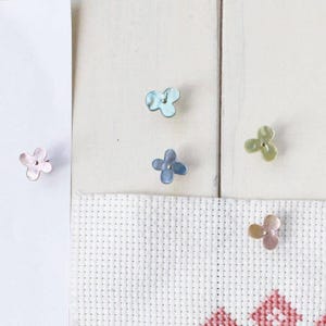以下が含まれることがあります： ピンク、ブルー、グリーンなどのパステルカラーの、花型のボタンが5つ。白い表面に配置され、クロスステッチの生地の一部が見えています。