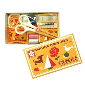 May include: A sewing kit with a yellow and white box, scissors, needles, thread, and a pin cushion. The box has the text "SAKURA CRAY-PAS" and illustrations of a dog, rose, and bird. The scissors are white.
