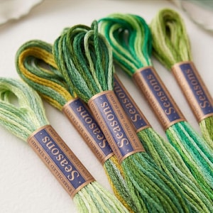 Pode incluir: Close-up de meadas de linha de bordar verde da coleção Seasons. A linha de algodão de seis fios apresenta várias tonalidades de verde. Cada meada é rotulada com a palavra "Seasons". O texto "Seasons 8000" está na parte inferior da imagem.