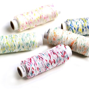 May include: A collection of colourful thread spools. The thread is white with flecks of pink, yellow, blue, and orange. The spools are arranged on a white surface. The thread is likely used for crafting or sewing projects. The spools have the word "hidama" printed on them.