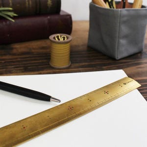 May include: A brass ruler with red dot accents and the text "O Cobans" rests on white paper, next to a black pen. A spool of yellow thread and a gray container with tools are in the background. The scene is set on a wooden surface.