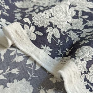 May include: A close-up shot of a fabric with a floral design. The fabric has a dark blue base with white floral patterns, including roses and other flowers. A white, textured border frames the fabric, providing a contrasting element to the design.