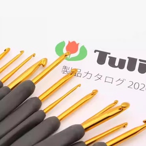Puede incluir: Primer plano de ganchillos con varillas de metal dorado y mangos grises oscuros. Los ganchillos están dispuestos en fila, con las puntas hacia arriba. La imagen también muestra un logotipo y texto japonés.