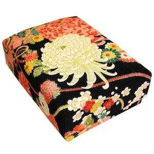 Puede incluir: Una caja rectangular de tela negra con un diseño floral. La caja presenta un gran crisantemo color crema y flores más pequeñas en rojo, amarillo y blanco. Los bordes están adornados con detalles rojos y dorados.