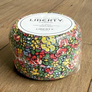 May include: A round bundle of Liberty Fabric yarn, wrapped in clear plastic. The fabric features a colorful floral pattern with red, yellow, and blue flowers on a dark green background. The label reads "YARN MADE FROM LIBERTY. FABRIC".