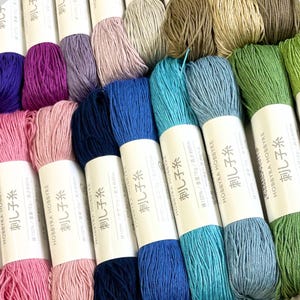 Può includere: Una collezione di matassine di filo da ricamo colorate in varie tonalità, tra cui rosa, blu, verde e viola. Ogni matassina è avvolta con un'etichetta bianca con testo giapponese. Il filo è ben avvolto, ideale per ricamo o altri lavori artigianali.
