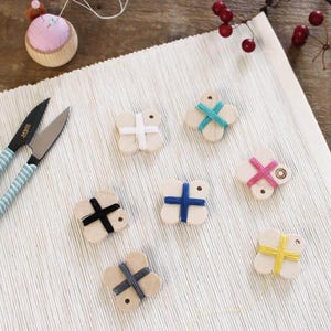 May include: A collection of wooden thread bobbins in various colors, including white, blue, pink, black, yellow, and gray. A pink pincushion and a pair of scissors with blue handles are also present. The items are arranged on a white textured surface.