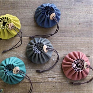 Puede incluir: Cinco pequeñas bolsas redondas de cuero con cordón en varios colores: amarillo, verde azulado, gris, rosa y azul. Cada bolsa tiene un cierre de botón de madera.