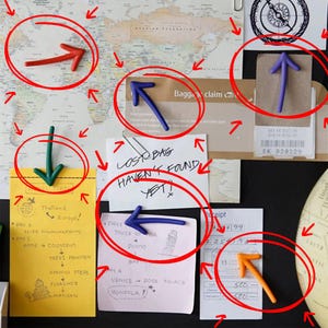 May include: A corkboard with travel-themed notes and arrow-shaped magnets. Notes include travel itineraries for Thailand and Europe, a baggage claim ticket, and a receipt. The text "LOST BAG HAVEN'T FOUND YET!" is visible.