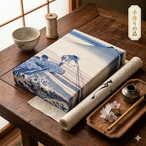 May include: A wooden table displays a wrapped package with a blue and white wave design, a rolled scroll, a small bowl, and a wooden tray with a brush and inkwell. The package is wrapped in a traditional Japanese style.