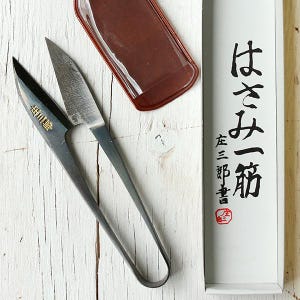 Puede incluir: Tijeras de costura japonesas con un estuche de cuero marrón y una caja blanca con texto japonés. Las tijeras son negras y plateadas, con un mango curvo y cuchillas afiladas. El estuche es rectangular.