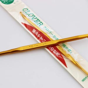 May include: Two gold-colored crochet hooks are displayed diagonally across a white and red package. The package has the word "CLOVER" and Japanese text. The crochet hooks are made of metal and have a smooth, shiny finish.