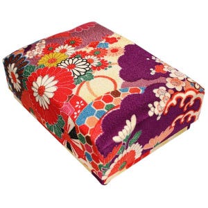 May include: A rectangular box with a vibrant floral pattern. The design features red, white, yellow, and purple flowers on a cream-colored background. The box has a soft, padded appearance, suggesting it may be used for storage or display.