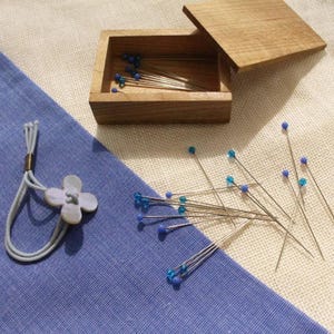 May include: A wooden box with a hinged lid, filled with sewing pins with blue and clear glass heads. A light blue flower-shaped button and a gray elastic loop with a gold-colored ring are on a blue fabric.