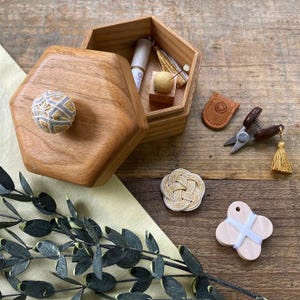 Peut inclure: Une boîte hexagonale en bois avec un bouton décoratif, ouverte pour révéler des accessoires de couture. Une petite paire de ciseaux, un étui en cuir et un pompon sont à côté de la boîte. Un nœud tissé et une forme en bois sont également visibles.