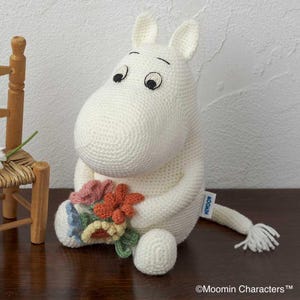 Könnte beinhalten: Gehäkeltes weißes Mumin-Plüschtier, das einen Strauß bunter Blumen hält. Das Plüschtier hat schwarz gestickte Augen und einen kleinen Schwanz. Das Mumin sitzt auf einer Holzoberfläche.