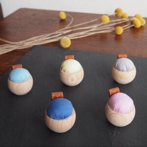 May include: Five wooden pincushions with different coloured fabric tops. The pincushions are arranged in a semi-circle on a black surface. The colours of the fabric tops are light blue, yellow, grey, pink, and dark blue.