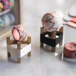 Puede incluir: Dos bolas Temari decorativas, una en forma de anillo y otra esférica, descansan sobre pequeños soportes de madera. La bola en forma de anillo presenta un colorido patrón geométrico, mientras que la bola esférica tiene una base blanca con costuras rosas y amarillas. Los soportes son dorados.