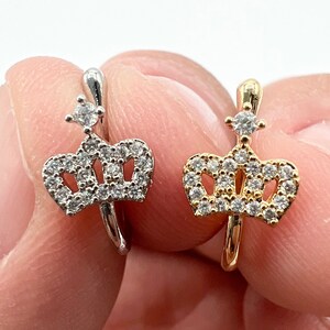 May include: Two crown rings, one silver and one gold, with clear cubic zirconia stones. The rings are on a finger.