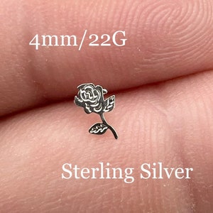 May include: A small silver rose flower stud earring, 4mm in size, made of sterling silver. The earring is shown on a finger.