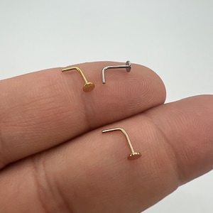 May include: Three nose studs in gold, silver, and rose gold. Each stud features a small, flat disc at the end of a curved post. The jewelry is displayed against a neutral background, highlighting the different metal finishes.