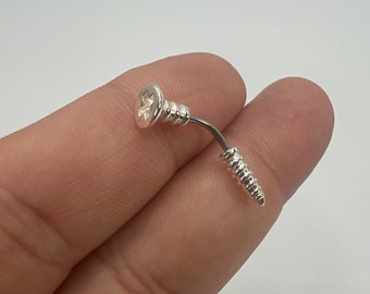 Puede incluir: Un pendiente plateado en forma de tornillo con un poste curvo. La cabeza del tornillo tiene un diseño en forma de estrella. El pendiente se muestra sobre un fondo claro.
