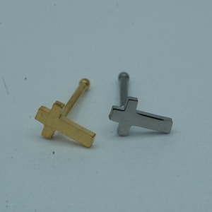 Op de afbeelding: Twee neus studs met kruisontwerpen. Eén is goudkleurig en de andere zilverkleurig. De studs hebben een rechte staaf met een kleine bal aan één uiteinde en een kruis aan de andere kant. De achtergrond is licht en neutraal.
