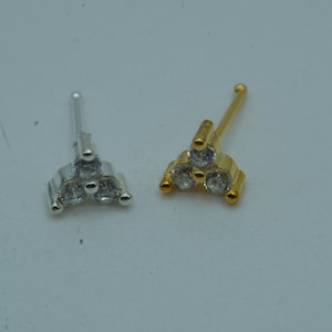 Könnte beinhalten: Zwei Nasenringe mit einem dreieckigen Design mit drei klaren Steinen. Ein Ring ist silber und der andere ist gold.